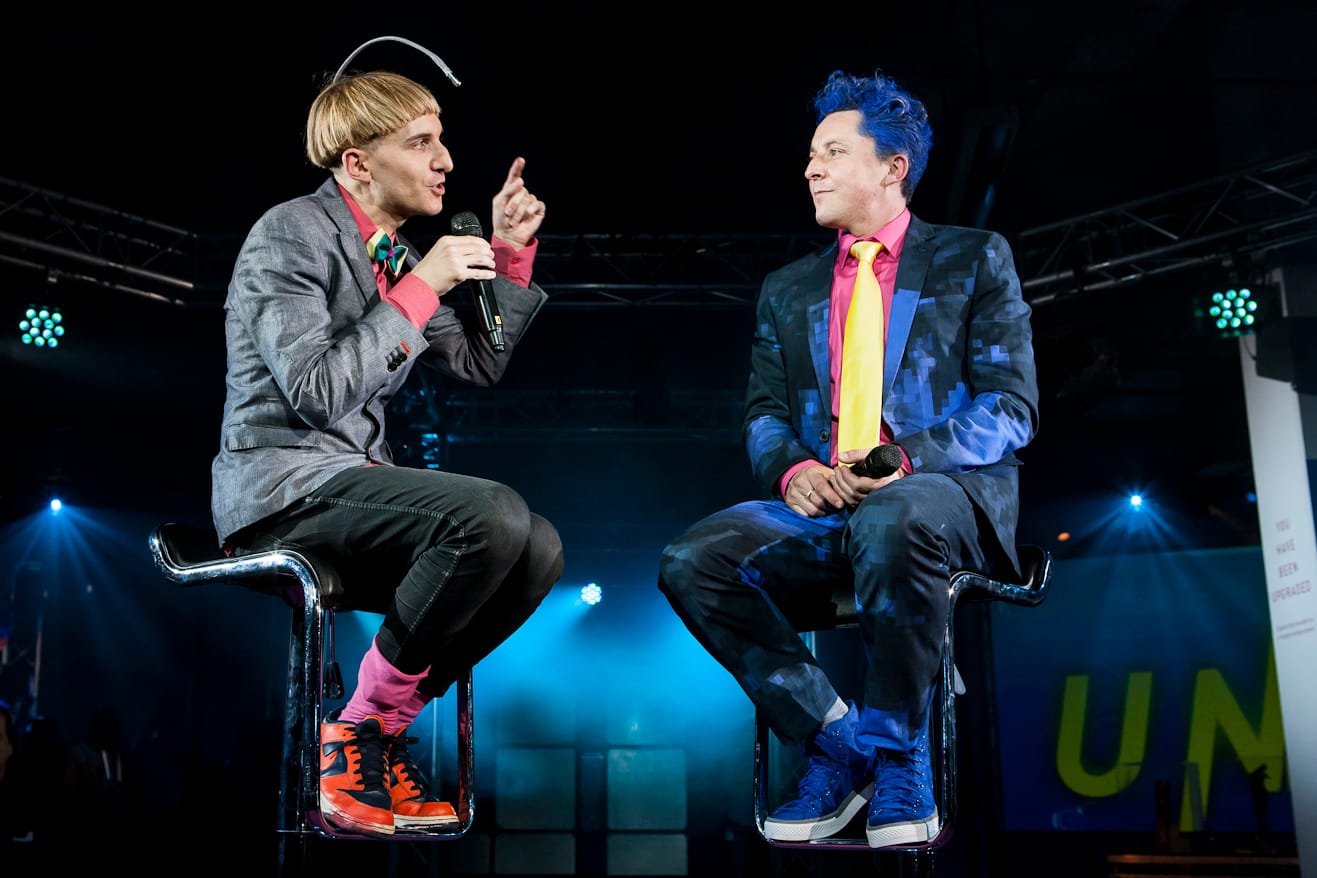 Two people sitting on a stage, one has an antenna sticking out of his head.